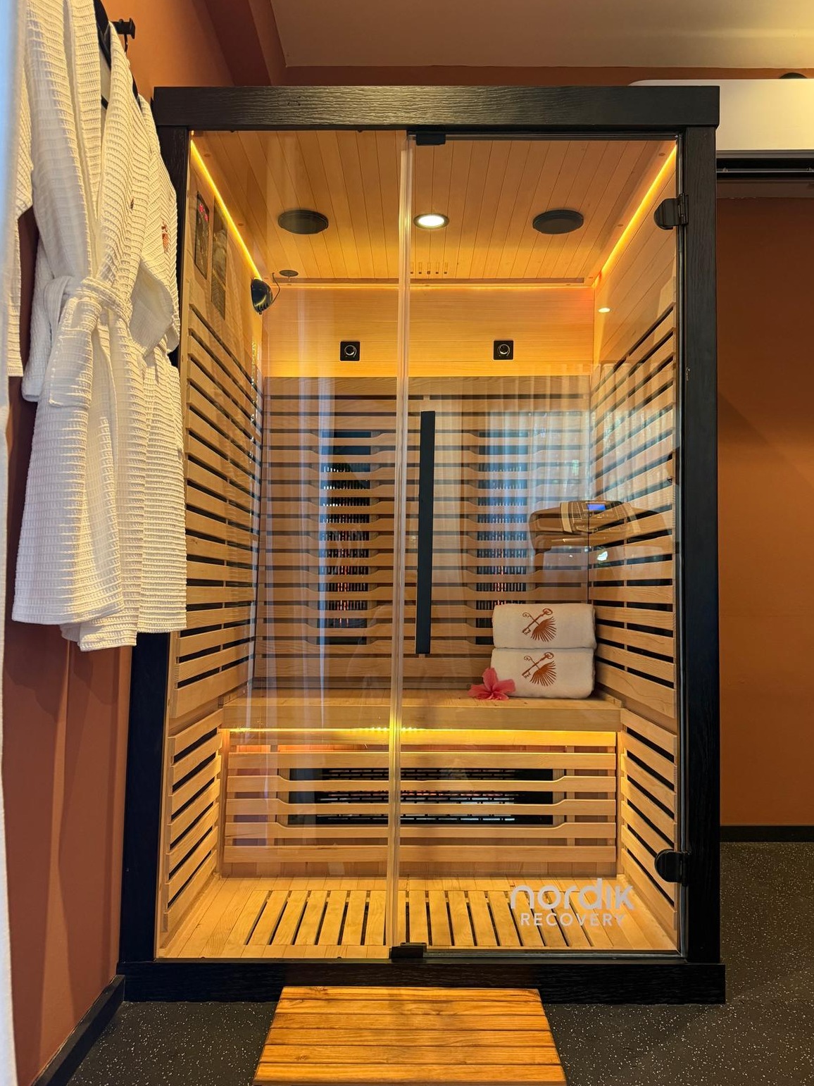 Infrared sauna cabin lit warm inside, rolled towels and a hibiscus flower on the bench, a teak mat at the door