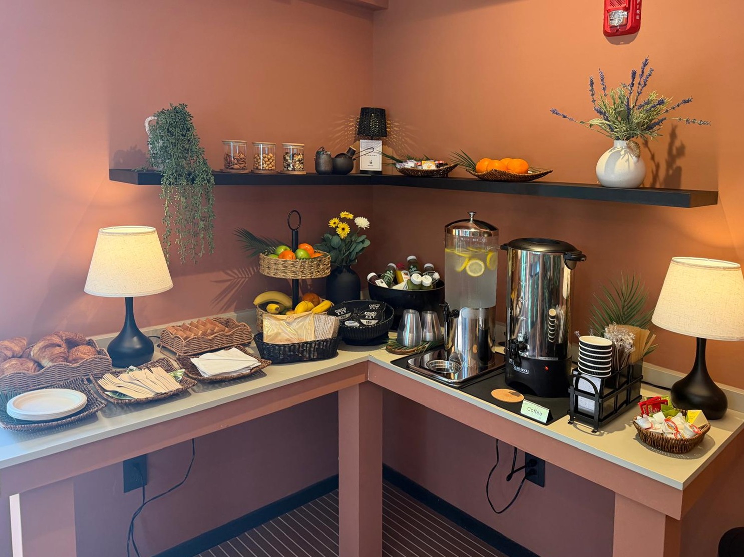 The Breakfast Table : croissants, présentoirs de fruits, jus sur glace, distributeur d'eau citronnée et urne à café sur un comptoir, lampes allumées contre des murs en terre cuite