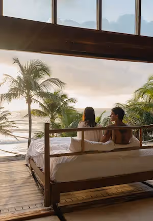 A couple sitting on the outdoor bed of a suite at La Valise Tulum, looking out at the beach and palms